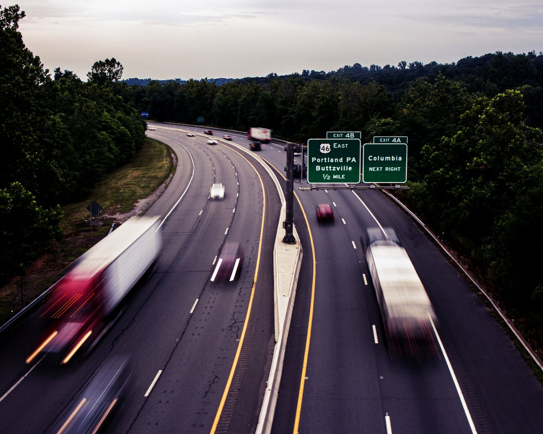 Traffic on a highway curving between trees, with an overhead direction sign